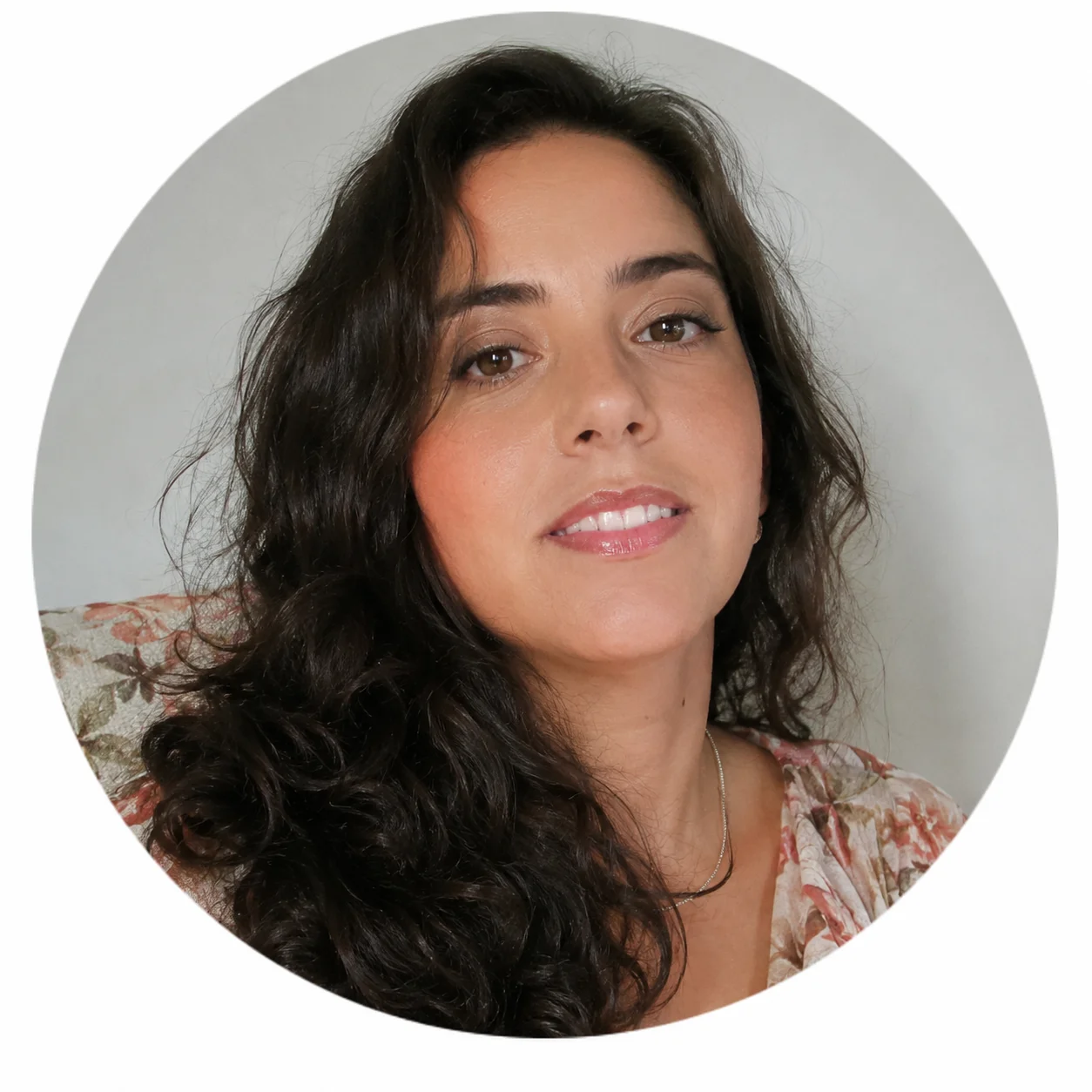 Retrato circular de mulher com cabelos longos e ondulados escuros, usando blusa floral em tons suaves e olhando para a câmera.